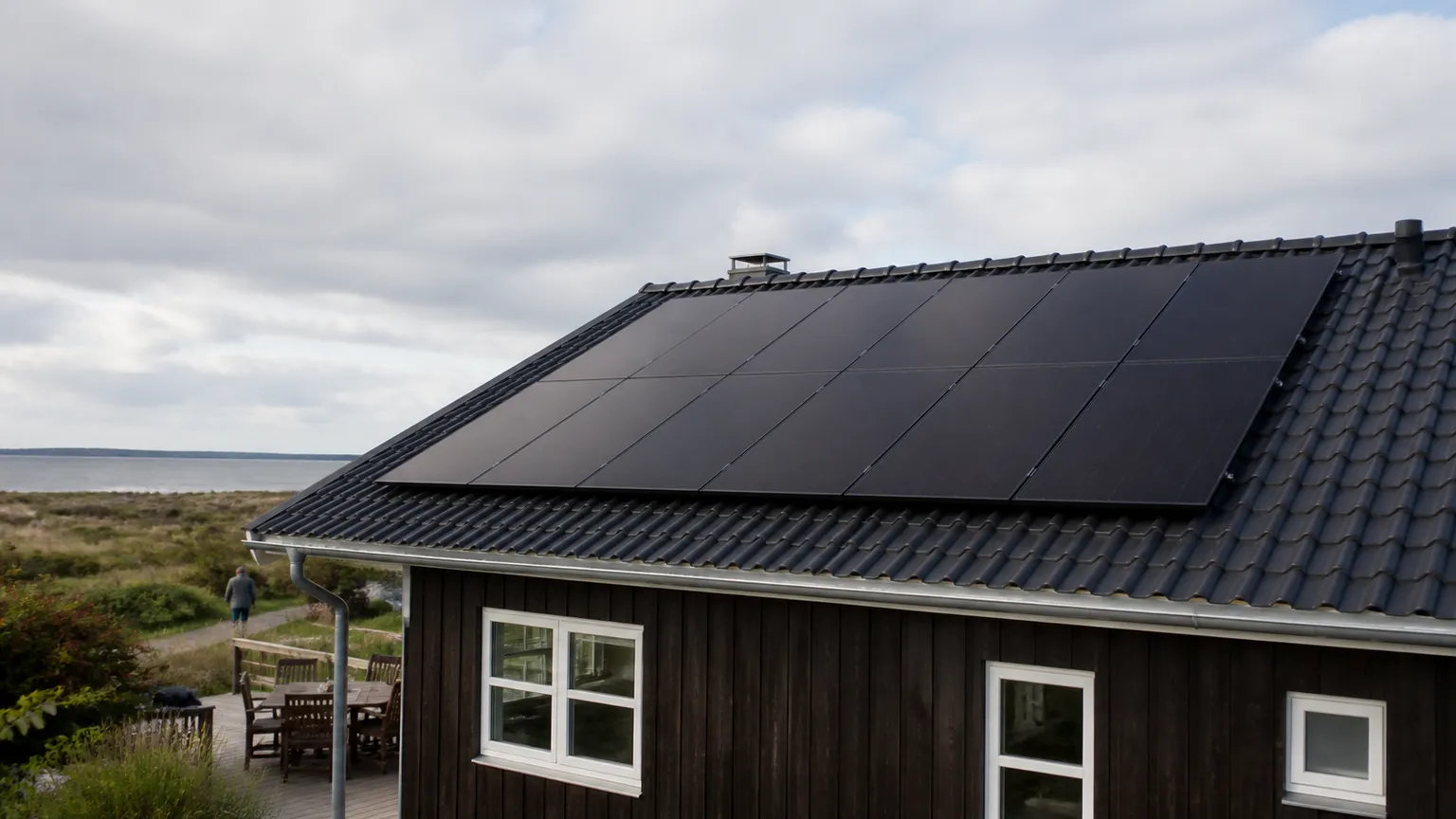Sorte solceller på taget af et dansk sommerhus ved kysten under en let skyet himmel.