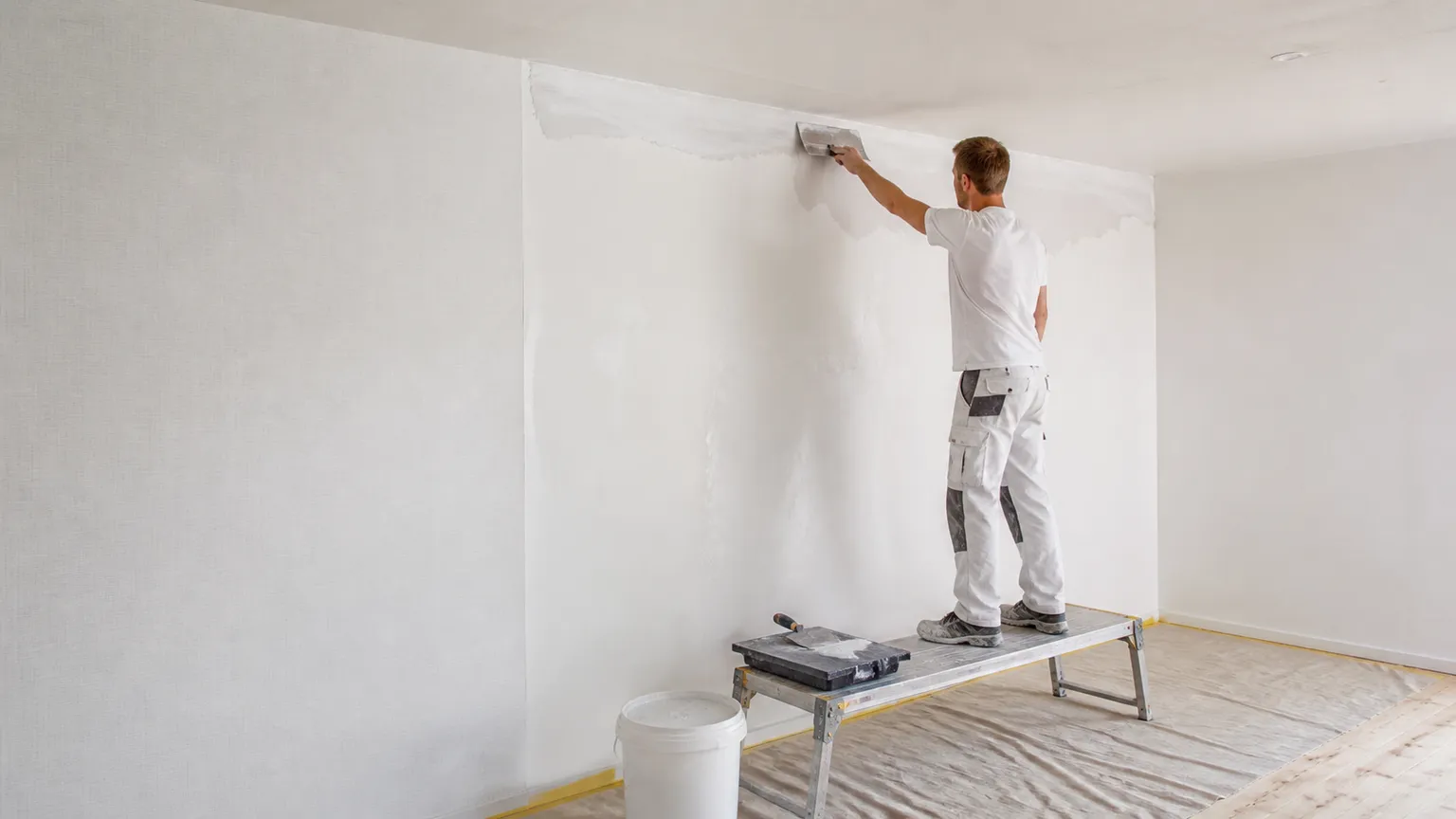Maler spartler en væg ved loftet i et lyst rum med filt, afdækket gulv og arbejdsplatform.