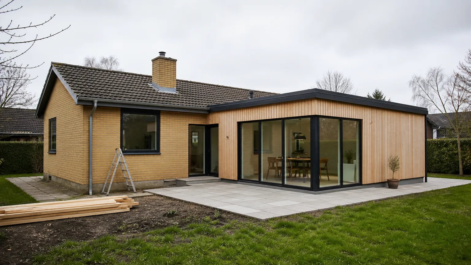Moderne tilbygning i træ og glas opført ved et dansk parcelhus i gule mursten fra 1970'erne.